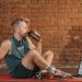Athletic man sitting on exercise mat with water bottle, taking a break from workout, in a rustic indoor setting.